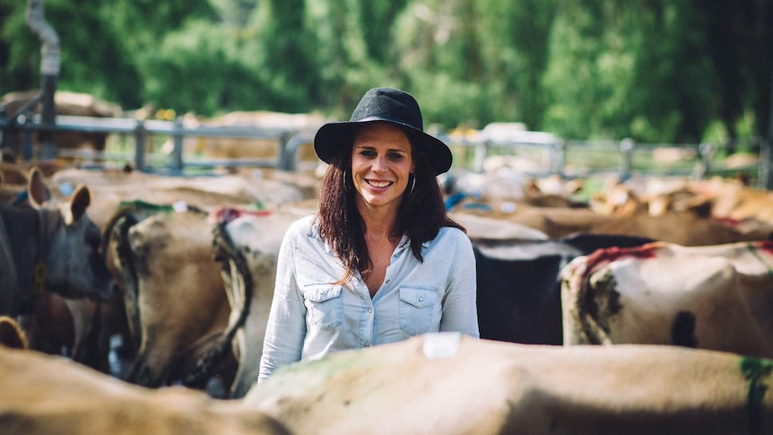 A woman with a broad-brim hat smiles as the camera surrounded by jersey cattle