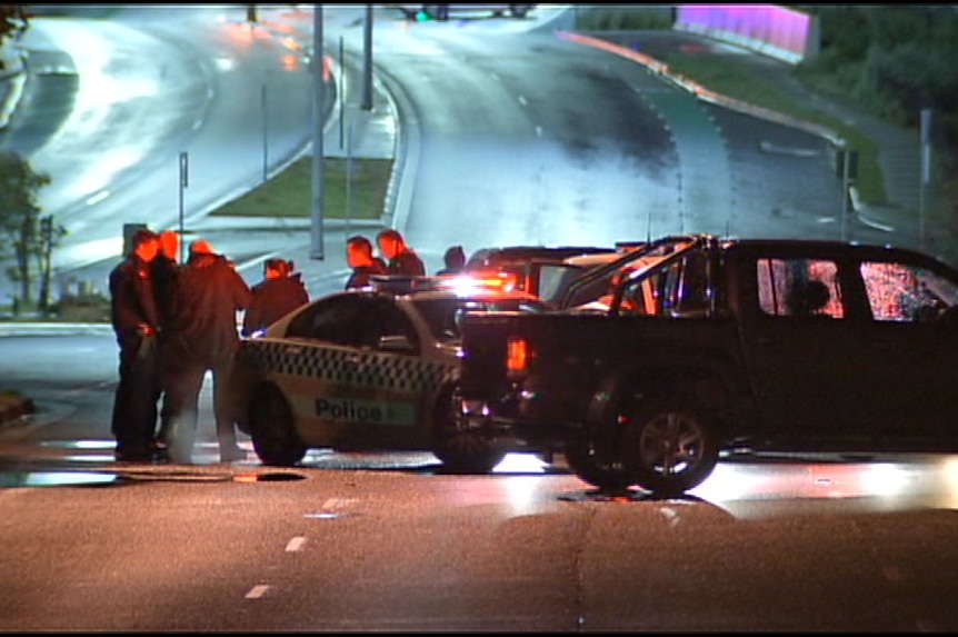 Police officers stand around a police car in the middle of the street at night, next to an abandoned 4WD.