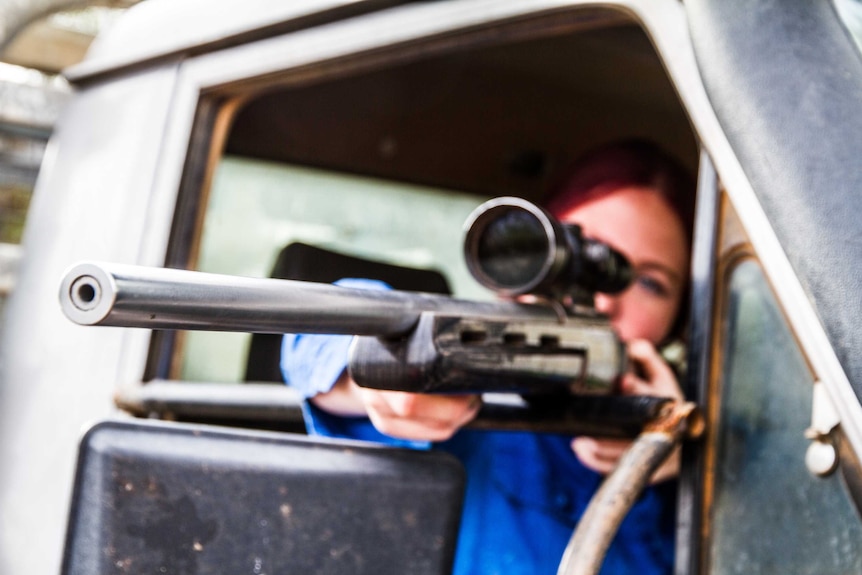 A woman in a blue shirt sits in the front of a vehicle and points a rifle out the window
