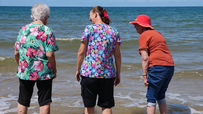 Two elderly women and a therapist standing in the shallow water of ocean with pants rolled up. 