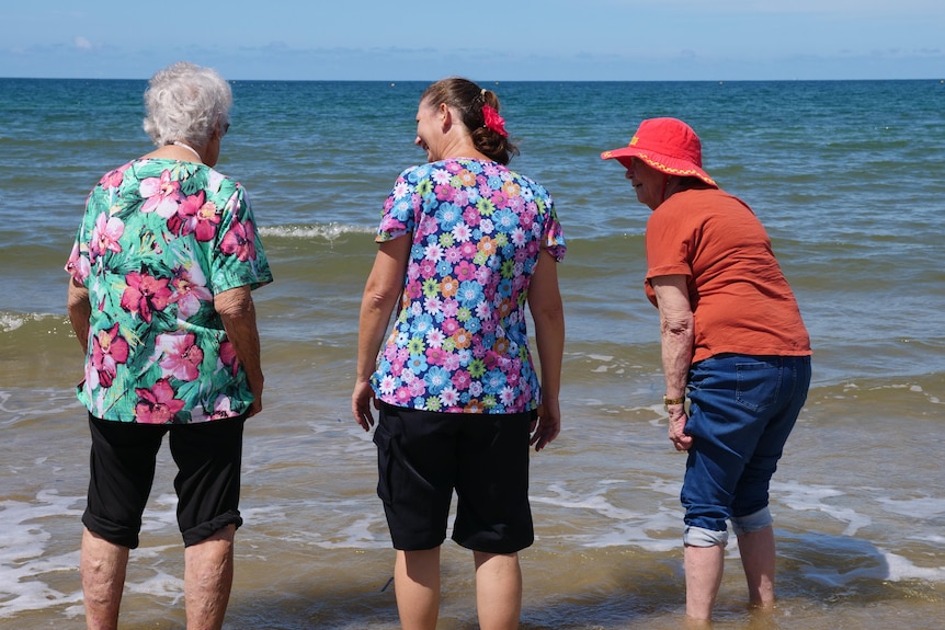 Two elderly women and a therapist standing in the shallow water of ocean with pants rolled up. 