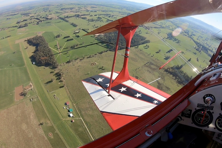 In the foreground is a red plane wing with white stripes and stars which is in the air, you can see a grassy landing strip below