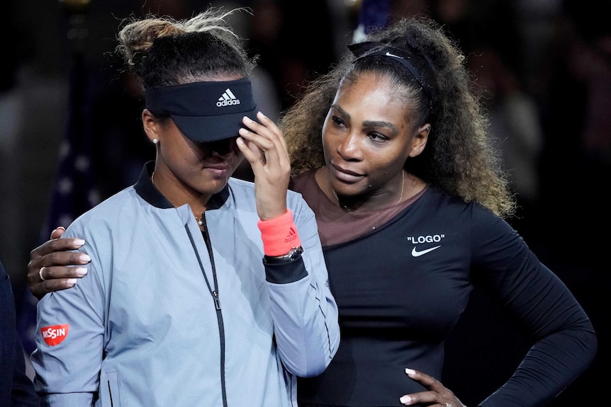 Naomi Osaka (left) bows her head as Serena Williams puts her arm around her.