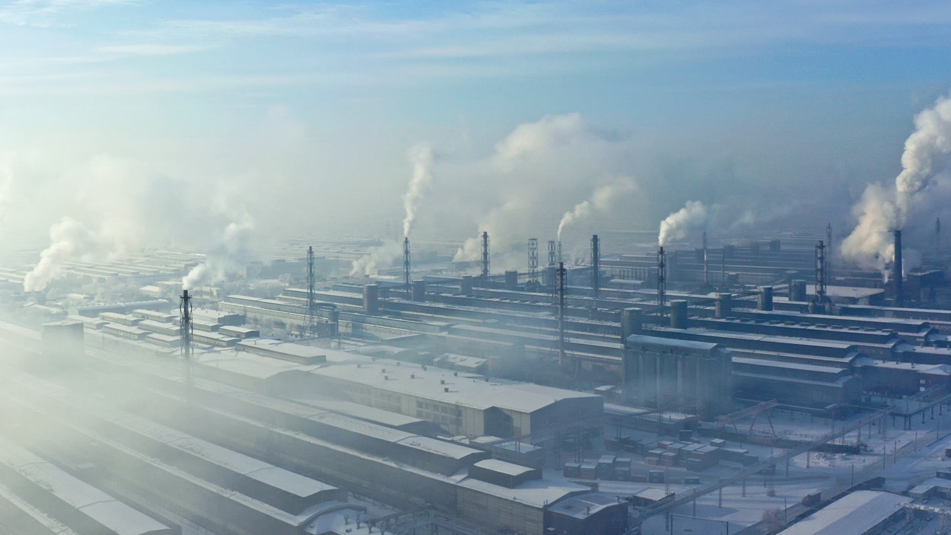 Factory complex on a snowy day. Chimney stacks across the complex billow smoke.