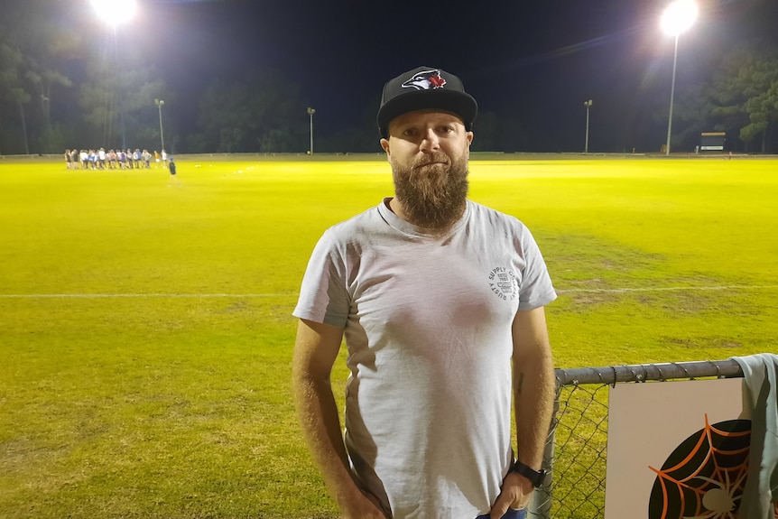 A man wearing a white T-shirt and a hat stands near a football oval under lights at night.