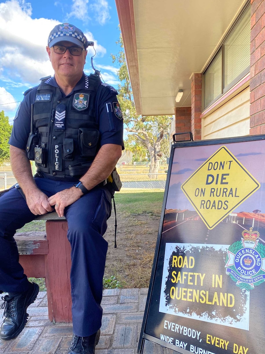 A man in police uniform sits on a bench next to a sign that says 'Don't Die on Rural Roads'.