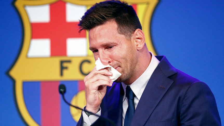Lionel Messi, wearing a suit and standing in front of a Barcelona logo, holds a tissue to his nose and cries