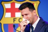 Lionel Messi, wearing a suit and standing in front of a Barcelona logo, holds a tissue to his nose and cries