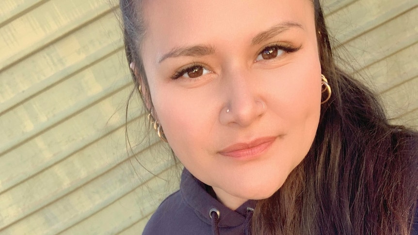 A woman with dark hair and earrings looks at the camera with a neutral expression