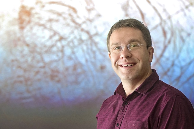 A man with short hair and glasses wearing a maroon shirt.