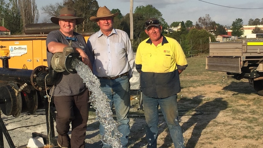 Three men stand next to a metal pipe that's spouting water. The men are smiling.