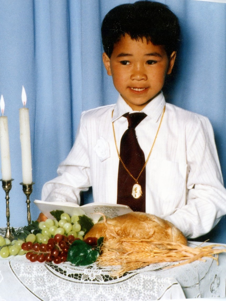 Young boy, wearing religious necklace, reading Christian book on a table with candles, bread and fruit.