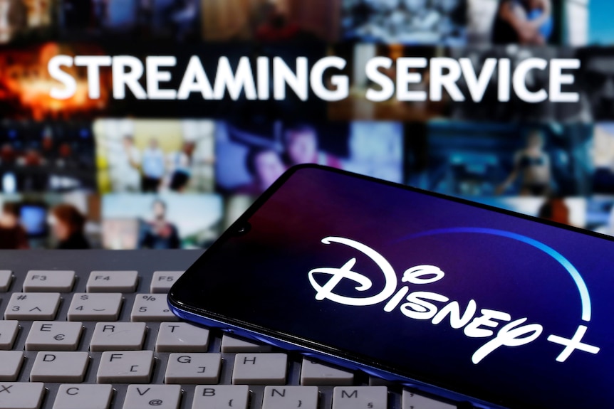 A smartphone with the Disney+ logo is seen on a keyboard in front of the words "Streaming Service".
