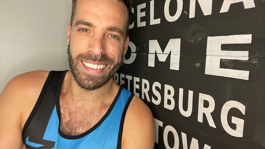 A man in a blue singlet stands in front of a black board with the names of cities.