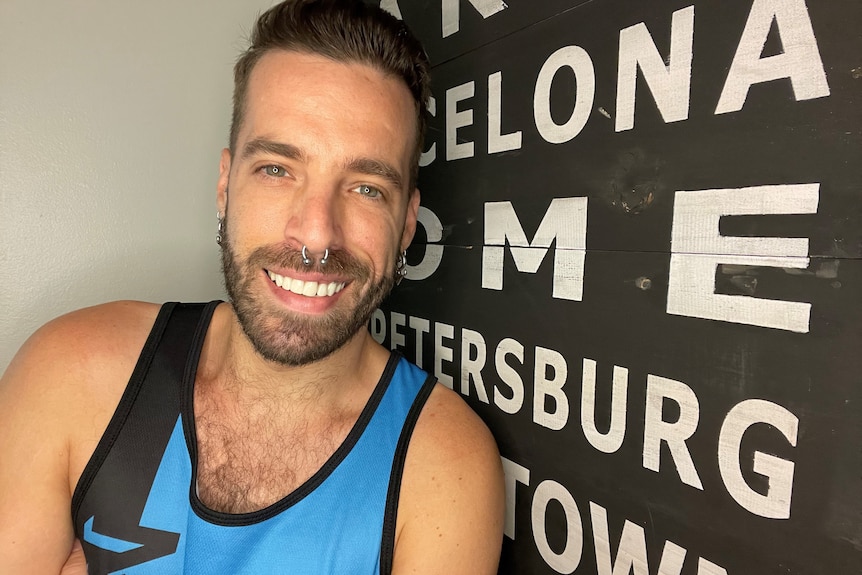 A man in a blue singlet stands in front of a black board with the names of cities.