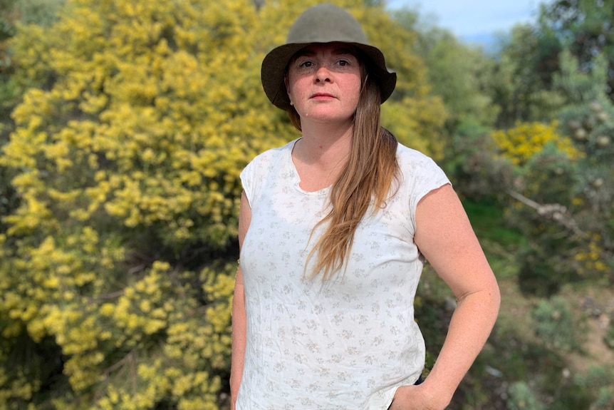 A woman stands outside wearing a broad-brim hat looking blankly past the camera in front of a yellow tree