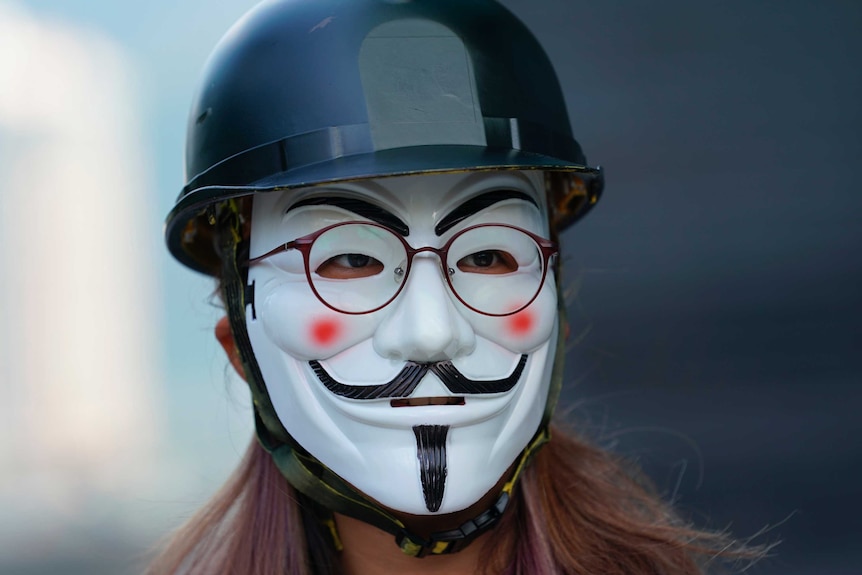 A student wears a Guy Fawkes mask as they march to a university. They have a helmet on, and glasses over the top.