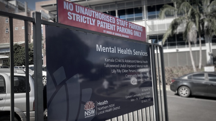 A large sign on a gate with 'mental health services'. building and carpark in background