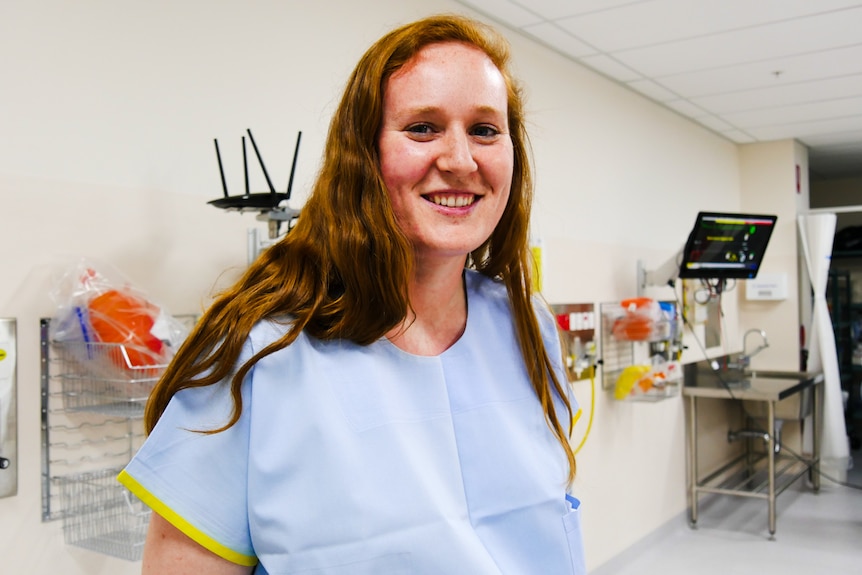 A girl smiles at the camera, wearing a light blue staff uniform, long red hair, hospital equipment behind.