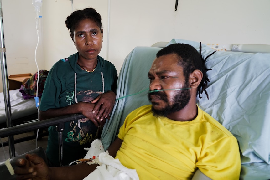 A woman in a green shirt stands next to a man in a hospital bed, both have pained expressions on their faces