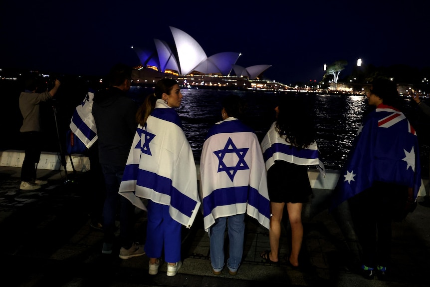 Young people watch the Opera House at night, wrapped in Israeli flags. The Opera House is lit up in blue and white
