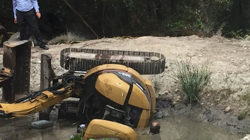Large excavator on side in muddy puddle. A man's head can be seen to the left as he freed from being trapped underneath.