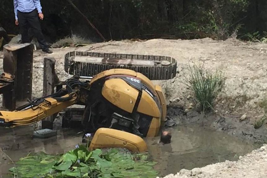 Large excavator on side in muddy puddle. A man's head can be seen to the left as he freed from being trapped underneath.