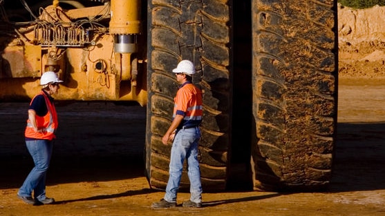 Two Macarthur Coal mining workers, with the back of a large coal mining truck towering over them.