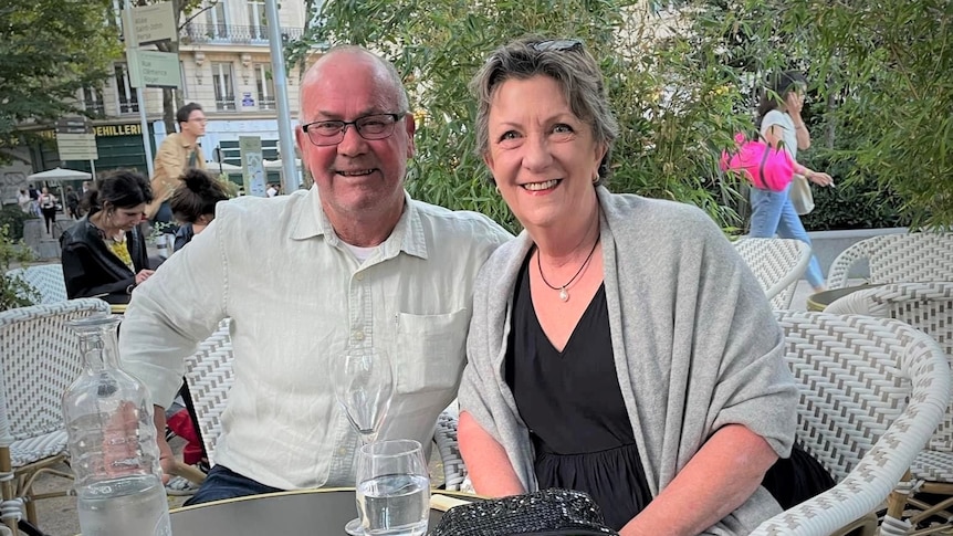 David and Maree Roberts sit at a cafe in France smiling at the camera. 