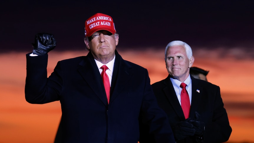 Donald Trump in a red cap pumps his first while Mike Pence looks on smiling. A deep orange sunset blooms behind them