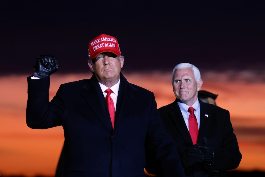 Donald Trump in a red cap pumps his first while Mike Pence looks on smiling. A deep orange sunset blooms behind them