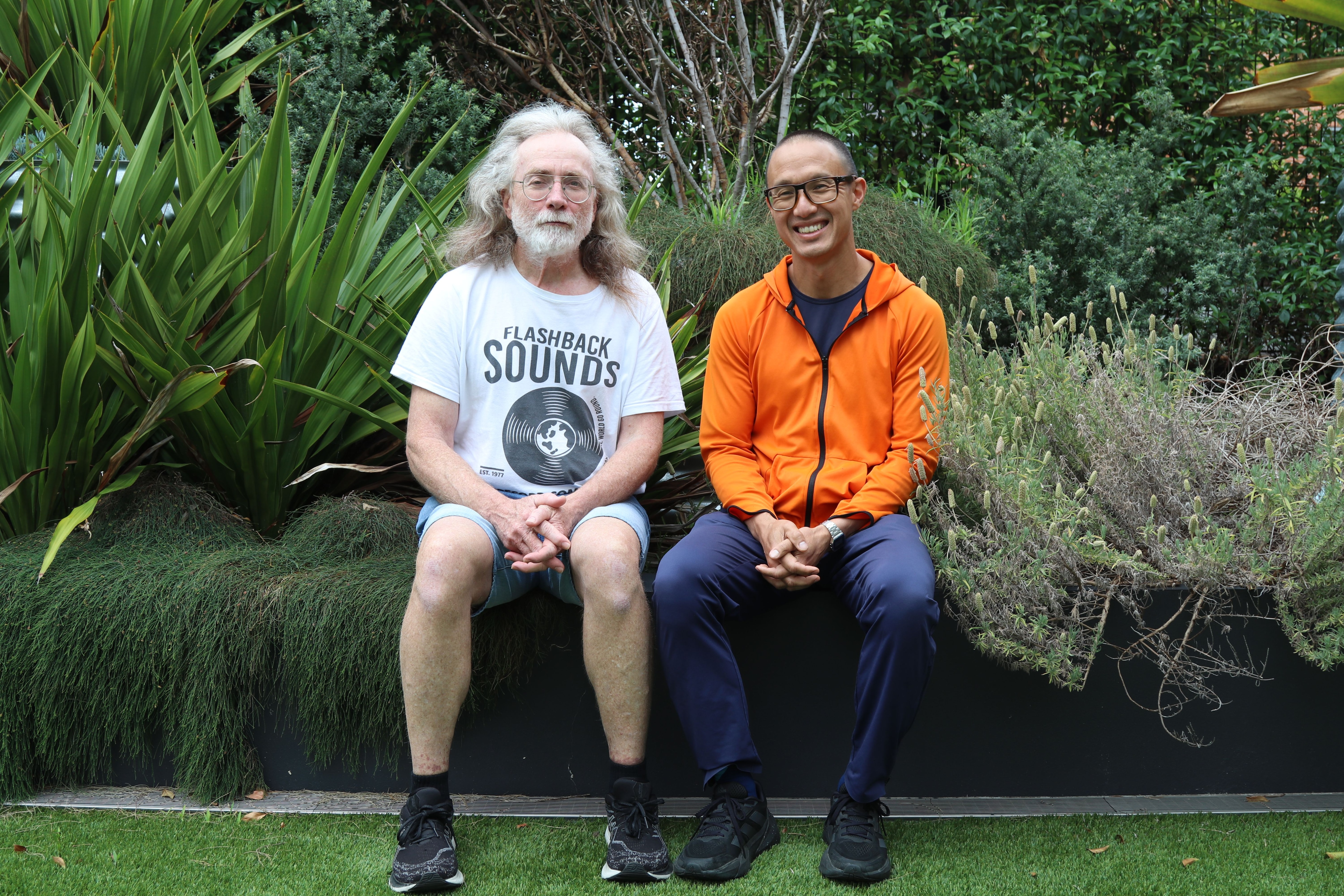 An older man with a silver beard and long grey hair sits in a garden next to a middle-aged man of Vietnamese background.