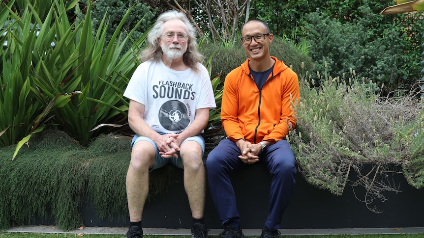 An older man with a silver beard and long grey hair sits in a garden next to a middle-aged man of Vietnamese background.