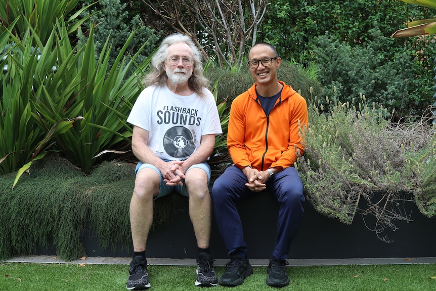 An older man with a silver beard and long grey hair sits in a garden next to a middle-aged man of Vietnamese background.