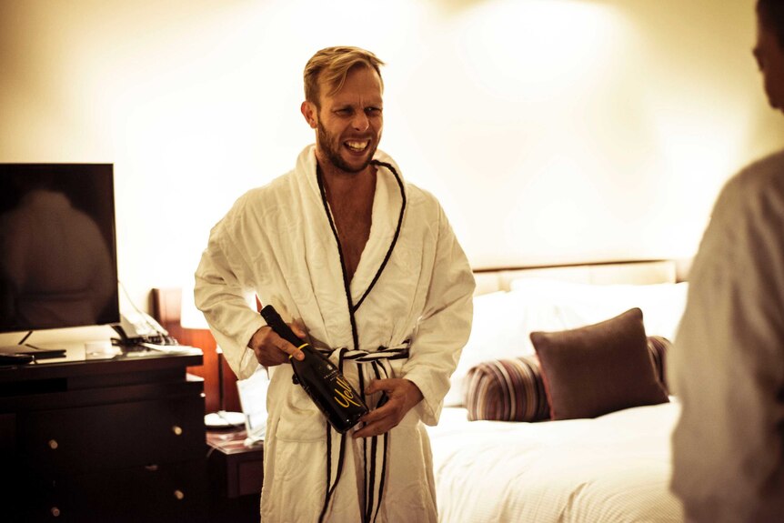 A man in a dressing gown smiling with a bottle of sparkling wine