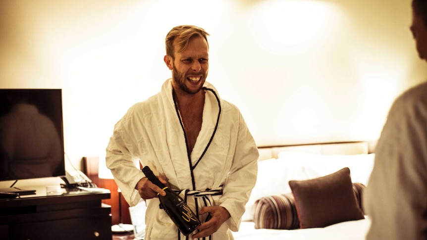 A man in a dressing gown smiling with a bottle of sparkling wine