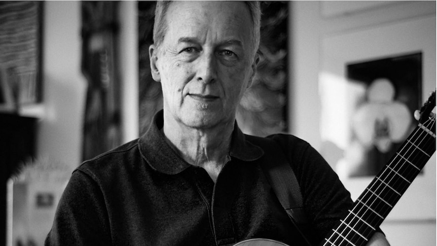 Black and white photo of Mike Rudd holding a guitar, wearing black shirt