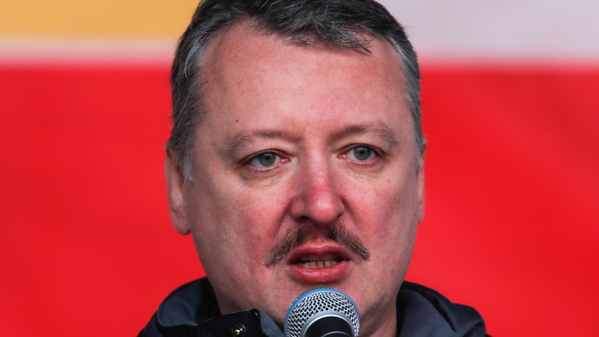 A close up of a man with a red tinge to his face and close cropped hair talking into a microphone against a red backdrop