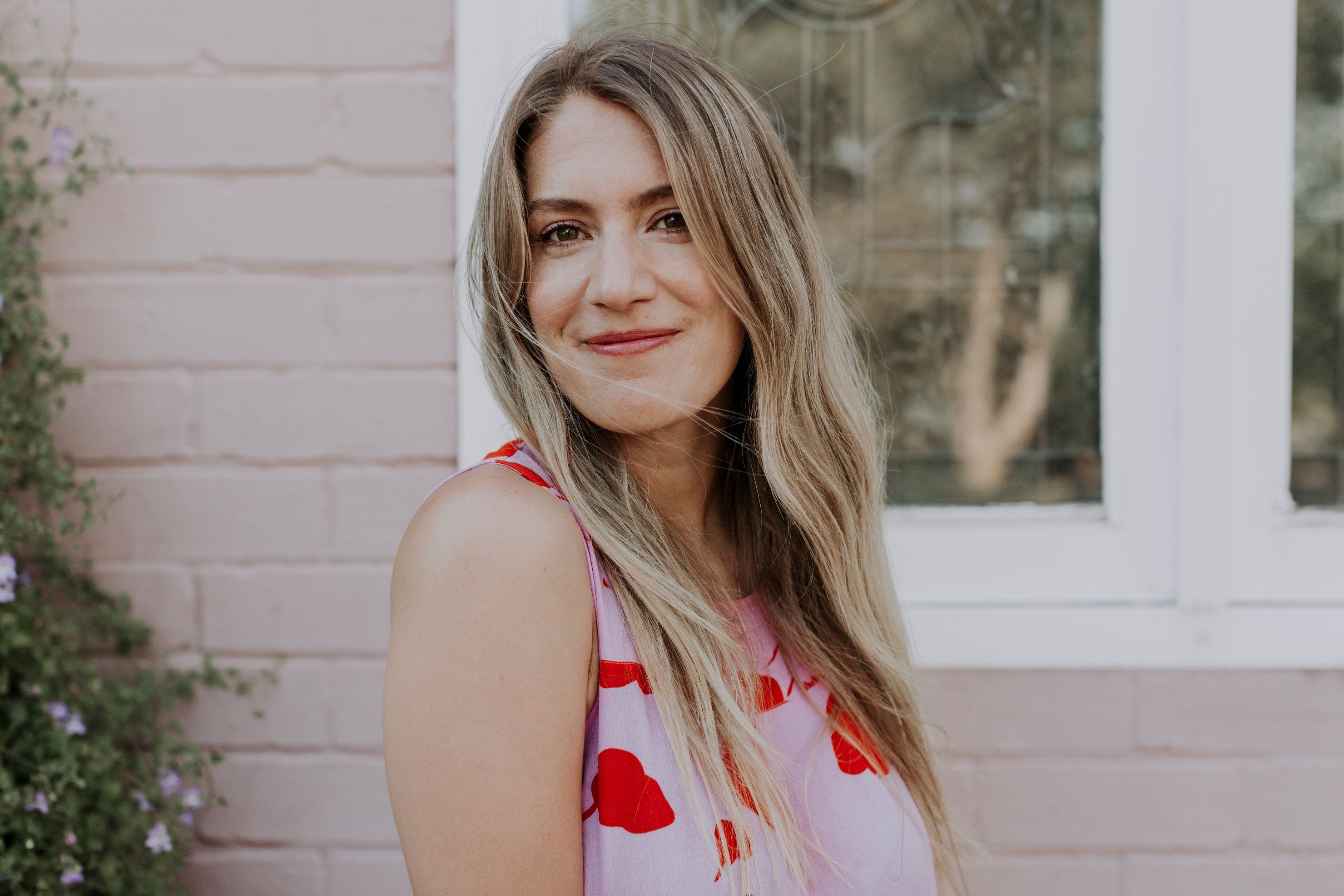 A close up of a smiling woman with long hair standing in front of a pink weatherboard building.