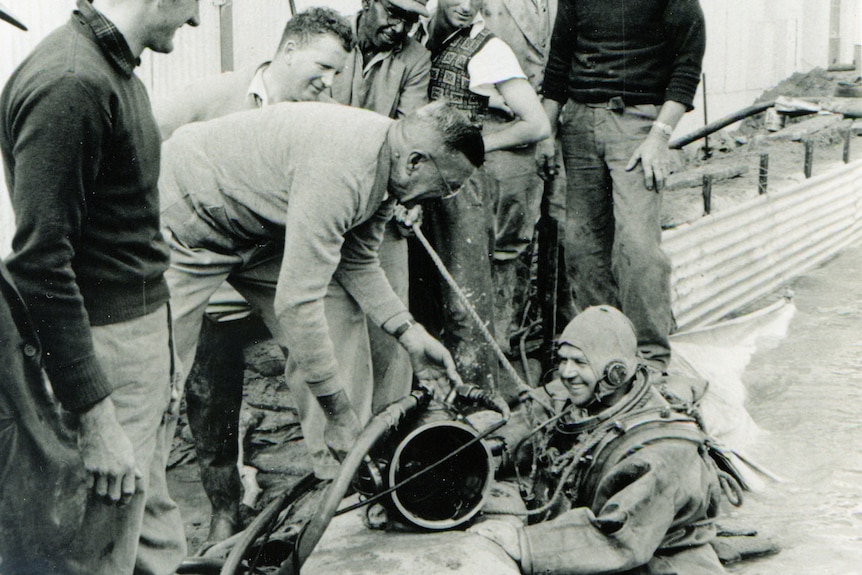 A man smiling, wearing a bathing cap, other men help him out of the water.