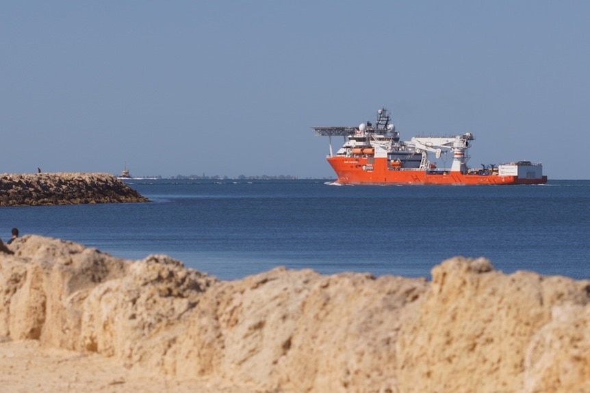 A red and white freight ship sails into port with a rocky groyne in the foreground