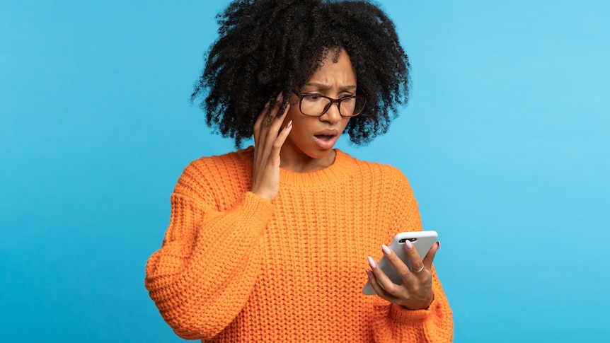 A young woman of African ethnicity wearing an orange jumper looking at a smartphone with opened mouth