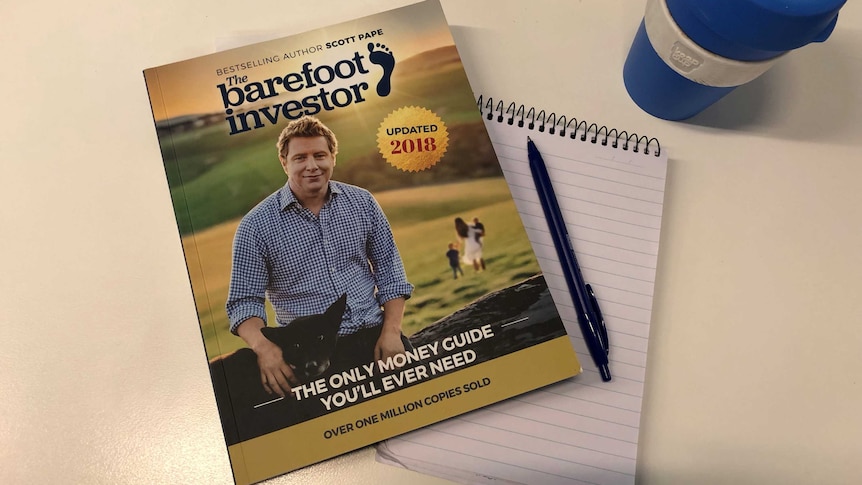 A coffee cup on desk with a copy of The Barefoot Investor and a notepad.