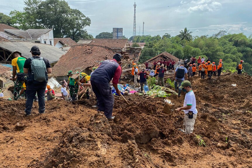 People dig through piles of mud while volunteers in orange vests gather in the distance