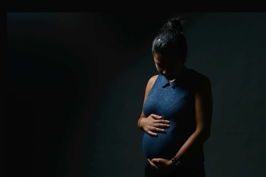 A woman with dark brown skin and face obscured in shadow wearing blue dress and holding her pregnant belly.