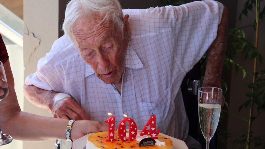 An elderly man in a checked shirt blows out candles reading 104 on a birthday cake.