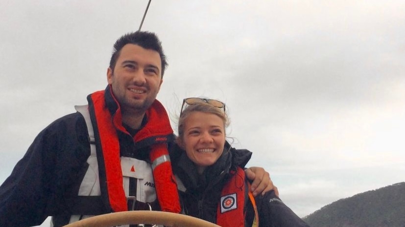 A bearded, dark-haired man embraces a fair-haired woman as they sail on a yacht