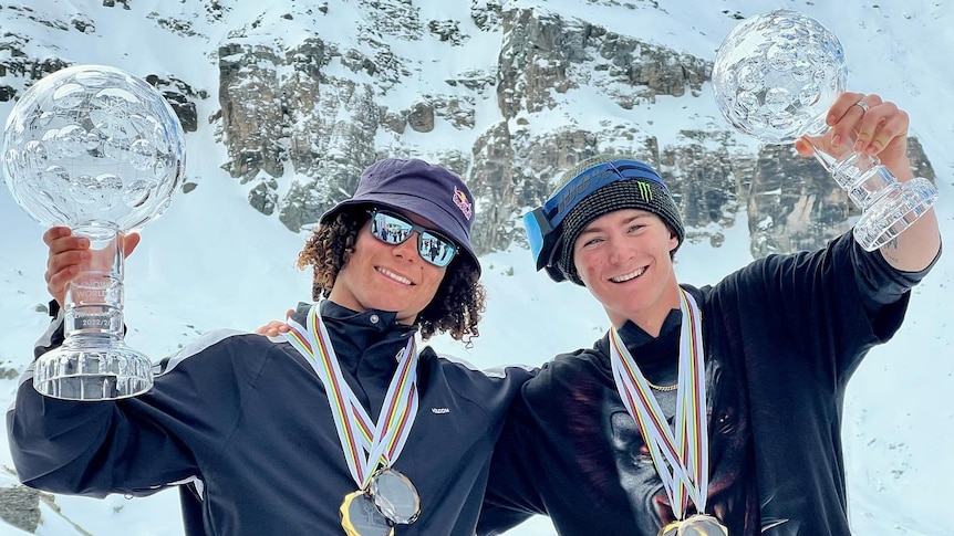 Two snowboarders with medals around their necks smile as they hold crystal globe trophies after the final event of a season.  