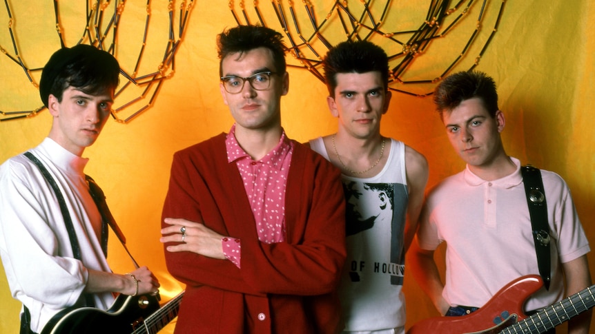 The four members of The Smiths post with their instruments backstage with cables hanging behind them on yellow wallpaper