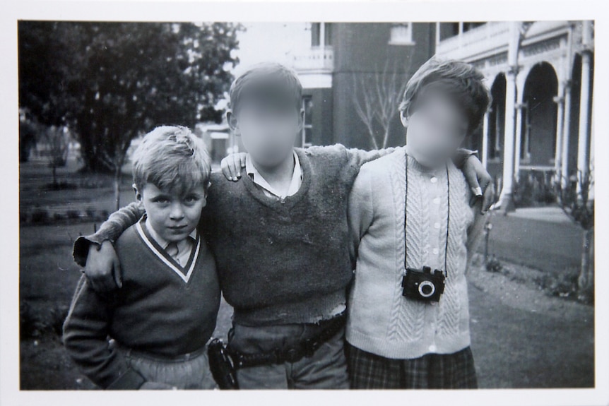 A black and white photo of three boys arm in arm - two of their faces have been blurred.